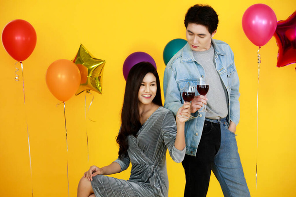 Studio shot of Asian happy young male and female lover couple model in fashionable wears sitting holding red wine glasses posing look at camera in celebration party with balloons on yellow background. - Photo, Image