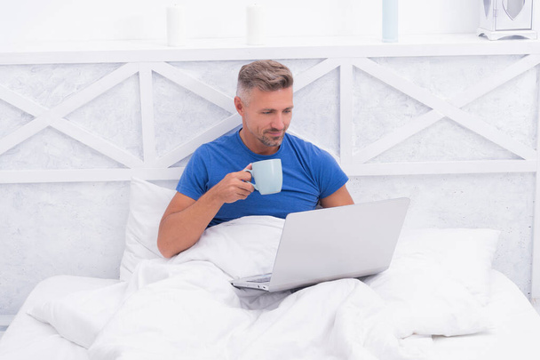 Happy guy relax drinking morning tea and using laptop computer in bed, relaxing - 写真・画像
