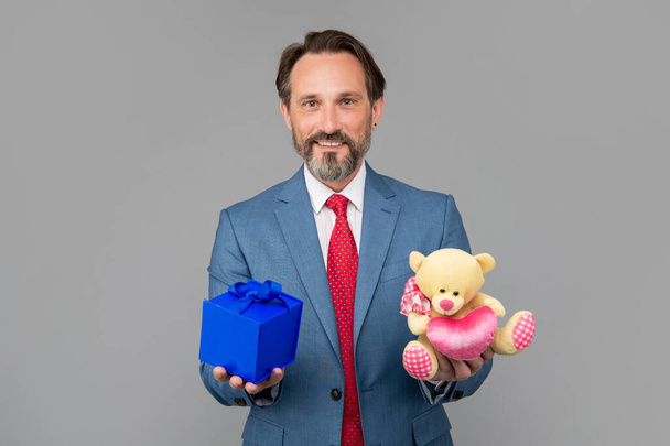 cheerful senior man in suit hold gift box and toy bear on grey background, love date - Valokuva, kuva