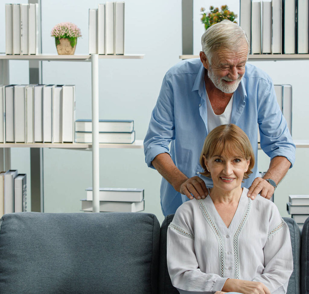 Caucasian old senior elderly gray bearded and hair male husband in casual wear standing behind massaging his lovely wife shoulder sitting smiling relaxing resting on sofa in living room at home. - Photo, Image