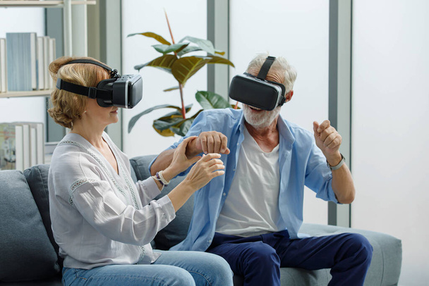 Caucasian old senior elderly grandparents couple wears virtual reality goggles headset. gray bearded and hair grandpa husband and grandma wife sitting together playing vr videogame in living room. - Fotó, kép