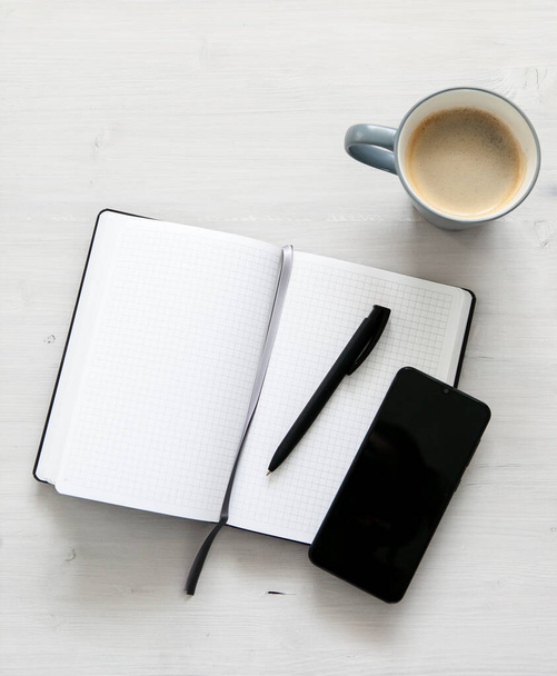 caderno branco aberto, um telefone celular preto e uma xícara de café no fundo de madeira leve - Foto, Imagem