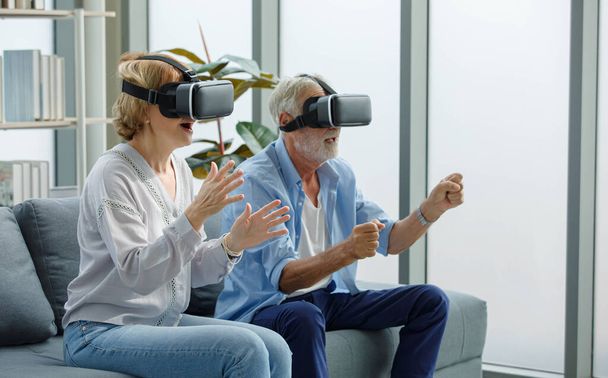 Caucasian old senior elderly grandparents couple wears virtual reality goggles headset. gray bearded and hair grandpa husband and grandma wife sitting together playing vr videogame in living room. - Photo, Image