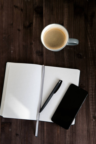 cuaderno blanco abierto, un teléfono móvil negro y una taza de café sobre un fondo de madera oscura - Foto, imagen