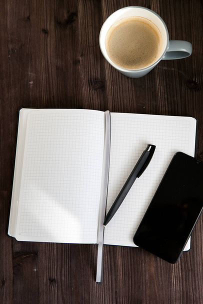 cuaderno blanco abierto, un teléfono móvil negro y una taza de café sobre un fondo de madera oscura - Foto, Imagen