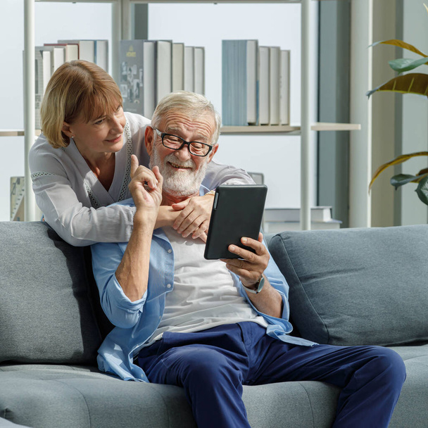 Vieux caucasien heureux grands-parents amoureux couple souriant en utilisant tablette ordinateur vidéo appel salutation en famille. Belle femme debout étreignant mari cheveux barbus gris assis sur le canapé. - Photo, image