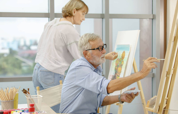 Caucasian old senior elderly male and female artist painter drawing painting flower bouquet picture with watercolor in palette and paintbrush on canvas paper on easel stand together in hobby workshop. - Foto, Imagen