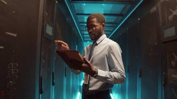 Colpo medio di giovane uomo afro-americano che indossa abiti formali utilizzando tablet digitale per la manutenzione del rack server cabinet - Filmati, video