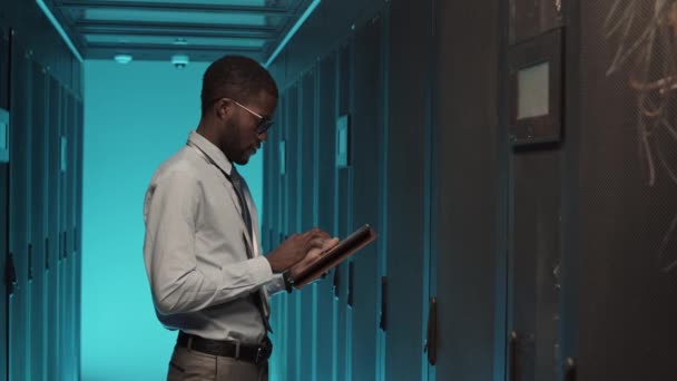 Foto media di un giovane afro-americano che indossa abiti formali in piedi al rack server cabinet e lavora su tablet digitale - Filmati, video