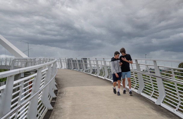 Ευρεία Shot of Two Teenage Boys Κοιτάζοντας ένα κινητό τηλέφωνο ενώ περπατάτε πάνω από μια γέφυρα σε μια πόλη με Copy Space - Φωτογραφία, εικόνα