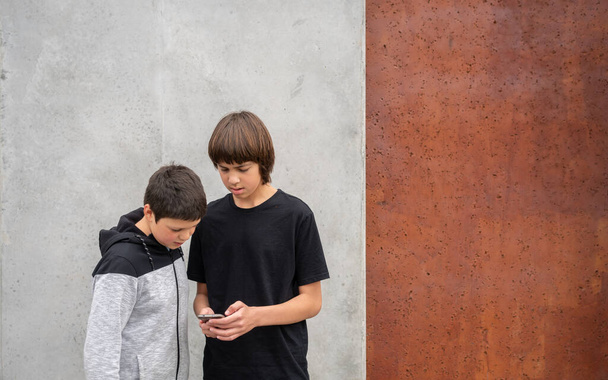 Deux adolescents regardant un téléphone portable debout devant un mur de béton dans un parc urbain avec espace de copie - Photo, image