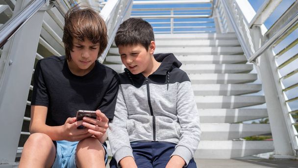 Κοντινό πλάνο δύο εφήβων αγοριών που κοιτάζουν ένα κινητό τηλέφωνο ενώ κάθονται σε σκαλοπάτια σε ένα πάρκο σε μια ηλιόλουστη μέρα - Φωτογραφία, εικόνα