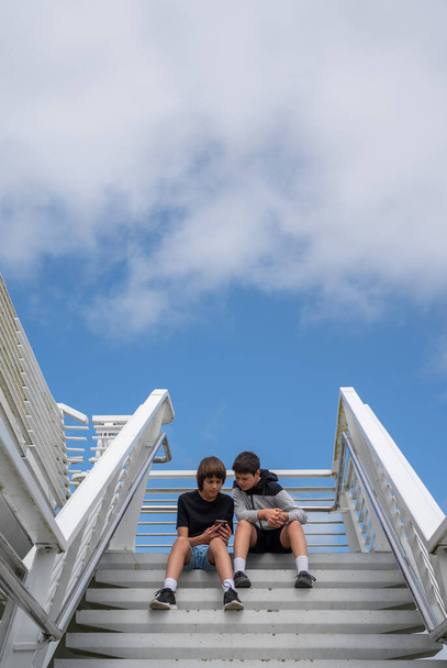 Широкий снимок двух мальчиков-подростков, смотрящих на мобильный телефон, сидя на ступеньках в парке с голубым небом и пространством для копирования - Фото, изображение