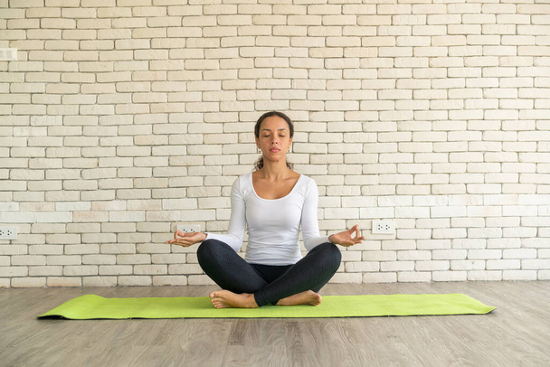 Latin woman practicing yoga on mat with white brick background - Foto, afbeelding