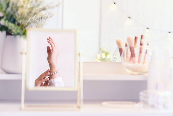 Reflection in table mirror of woman hand  in pearl bracelets on dressing table with make up accessories. Self-Care. White interior - Foto, afbeelding