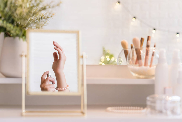 Reflection in table mirror of woman applying perfume on hand. Female party preparation. Christmas, light bulbs, eucalyptus, fir,  pine branches in a vase  on dressing table with make up accessories.  - Photo, Image