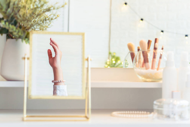 Reflection in table mirror of woman hand  in pearl bracelets on dressing table with make up accessories. Self-Care. White interior - Photo, Image