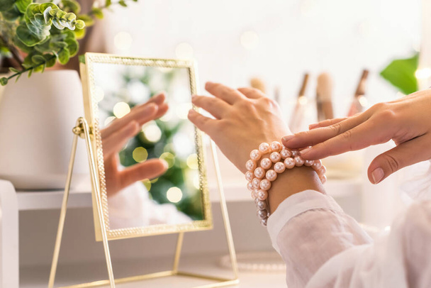 Woman puts on jewelry, pearl bracelets on the hand and  sitting near dressing table with make up accessories. Female party preparation. Reflection of christmas tree, light bulbs in table mirror. - Foto, immagini