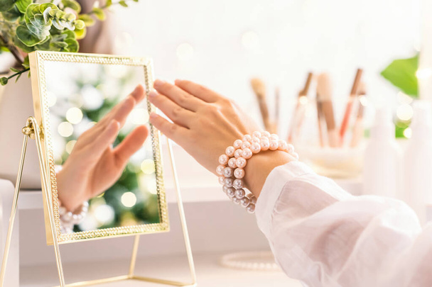 Woman hand in pearl bracelets sitting near dressing table with make up accessories. Reflection of christmas tree, light bulbs in table mirror. - Foto, Bild