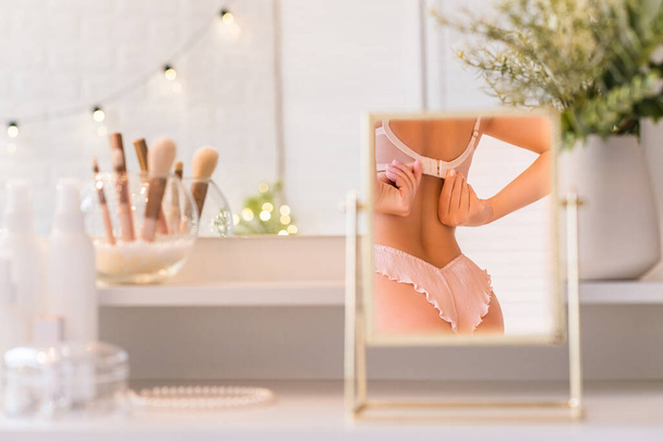 Reflection in table mirror of woman body in underwear. Female party preparation. Christmas, light bulbs, eucalyptus, fir,  pine branches in a vase  on dressing table with make up accessories.  - Fotografie, Obrázek