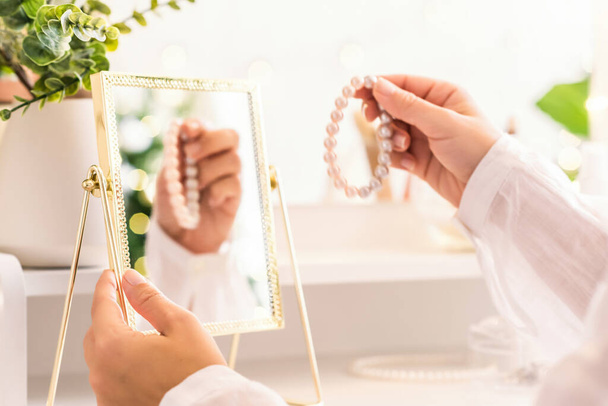 Woman puts on jewelry, pearl bracelets on the hand and  sitting near dressing table with make up accessories. Female party preparation. Reflection of christmas tree, light bulbs in table mirror. - Foto, Bild