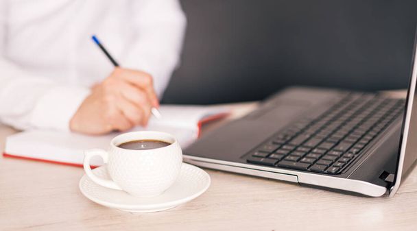 Mujer escribe con pluma en el cuaderno delante de la computadora portátil y la taza de café - Foto, Imagen