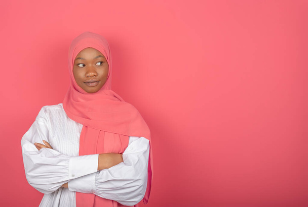 Ικανοποιημένο θρησκευτικό γυναικείο μοντέλο έχει τα χέρια διπλωμένα, κοιτάζει πέρα, φοράει ροζ μαντίλα στέκεται σε ροζ φόντο, αντίγραφο χώρο για κείμενο - Φωτογραφία, εικόνα