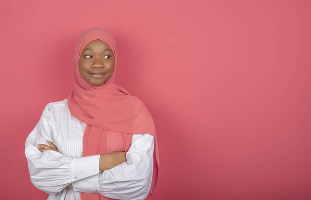 Ικανοποιημένο θρησκευτικό γυναικείο μοντέλο έχει τα χέρια διπλωμένα, κοιτάζει πέρα, φοράει ροζ μαντίλα στέκεται σε ροζ φόντο, αντίγραφο χώρο για κείμενο - Φωτογραφία, εικόνα