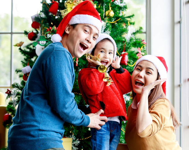 Happy Asian family father mother and little daughter girl wears sweater and red and white Santa Claus hat look at camera taking photo together in front decorated Christmas pine tree in living room. - Φωτογραφία, εικόνα