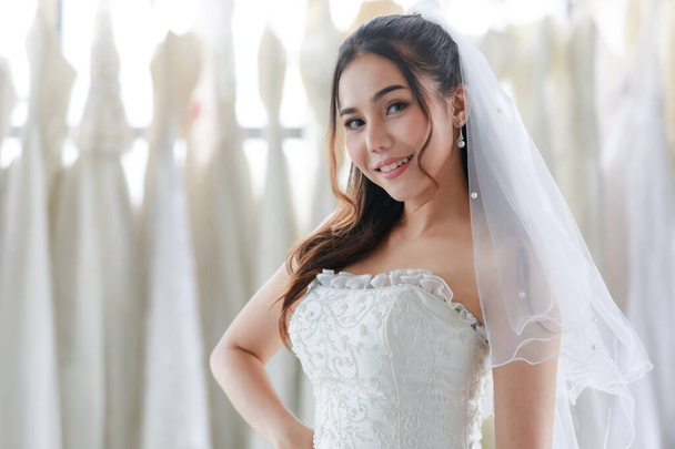 Primer plano retrato de la joven asiática hermosa novia pelo largo feliz en vestido de novia blanco con ver a través del velo de pie mirada sonriente a la cámara en el vestidor lleno de vestidos de fondo borroso. - Foto, imagen