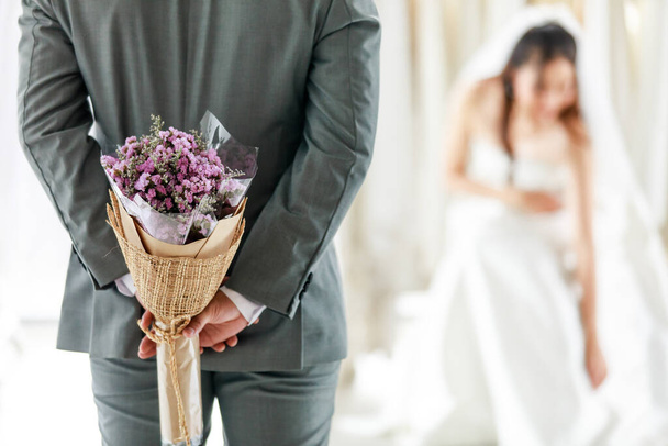 Asiatische junge schöne langhaarige Braut in weißem Brautkleid mit durchsichtigem Spitzenschleier sitzt lächelnd und wartet auf den Bräutigam im grauen Anzug, der den Blumenstrauß hinter dem Rücken versteckt, um in der Garderobe zu überraschen. - Foto, Bild