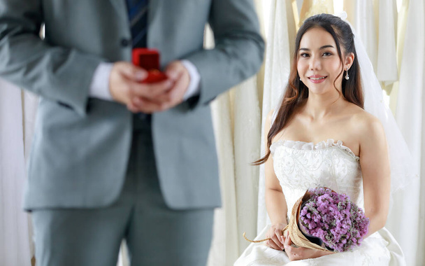 Felismerhetetlen vőlegény szürke formális öltöny stand hold piros doboz gyémánt gyűrű felkészülés ad ázsiai fiatal szép boldog menyasszony fehér esküvői ruha gazdaság virág csokor homályos háttérrel. - Fotó, kép