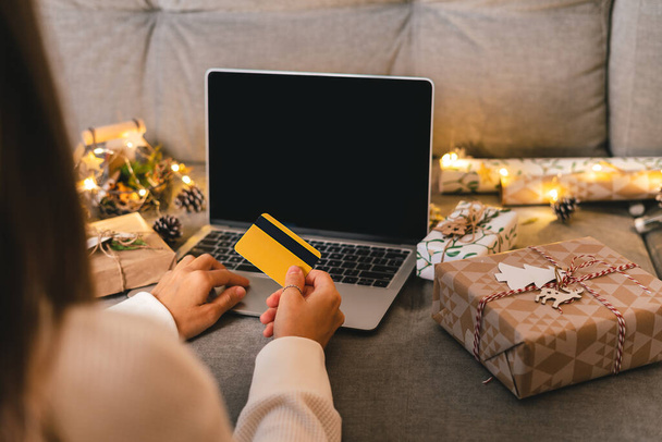 Close up of woman hands with credit card, gifts, and mock up screen laptop. Online shopping at Christmas holidays. Cropped female sit on couch with natural eco presents and decor. Merry Christmas - 写真・画像