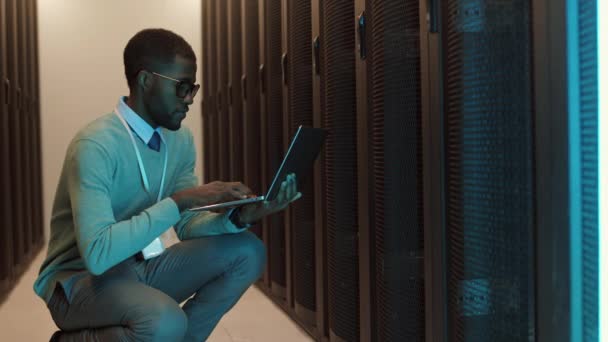 Genç Afrikalı erkek bilişim idarecisinin raf sunucusu dolabına çömelmiş ve bilgisayarla çalışmalarını analiz ederken orta boy bir fotoğrafı. - Video, Çekim