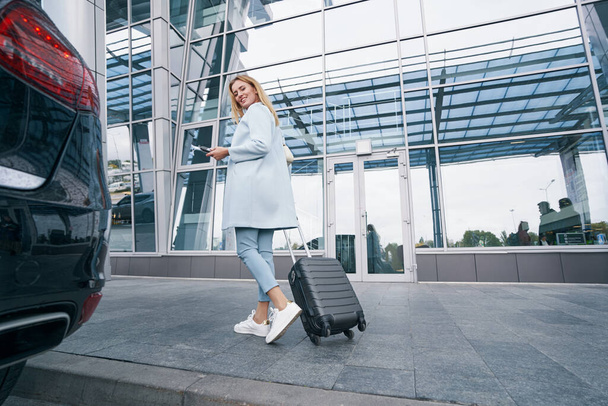 Joyous γυναίκα τουρίστρια με τρόλεϊ βαλίτσα στέκεται σε εξωτερικούς χώρους - Φωτογραφία, εικόνα