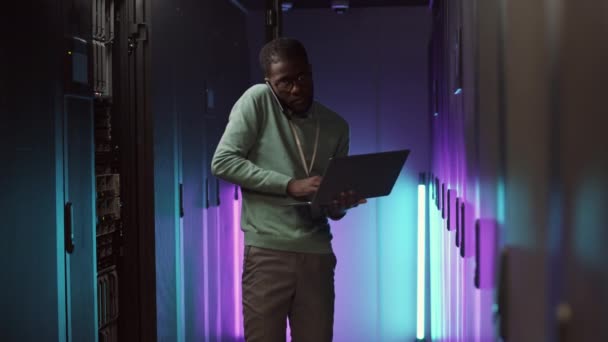 Colpo medio di giovane uomo afro-americano che analizza il lavoro di hardware nel data center utilizzando laptop mentre consulta il collega per telefono - Filmati, video