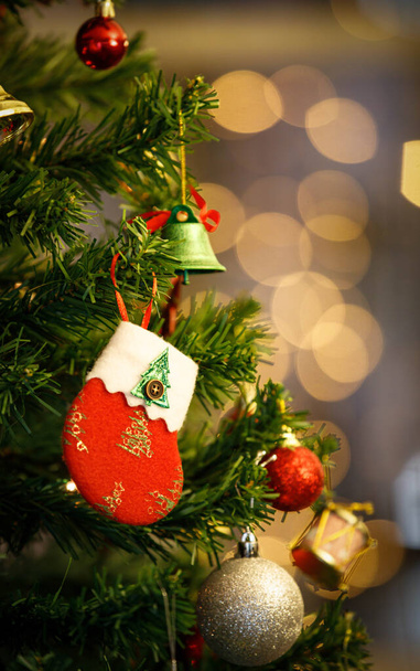 Kırmızı ve beyaz Noel arifesi çorabının yakın plan resmi yeşil Xmas çam ağacı dalında asılı, parlak bir sopa küresi ve zili ile önde bulanık altın rengi düşük ışık arka plan bokeh. - Fotoğraf, Görsel