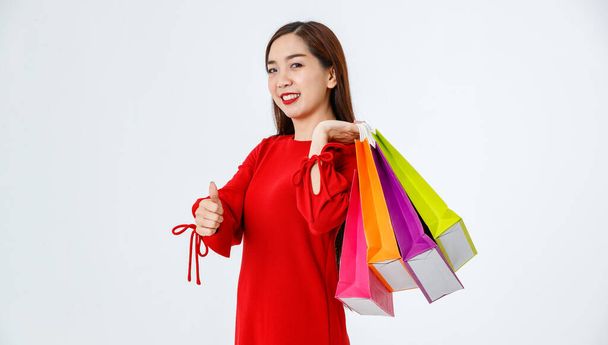 Calm Asian female customer with paper shopping bags touching lips and showing thumb up sign while looking at camera on white background in studio - Photo, Image