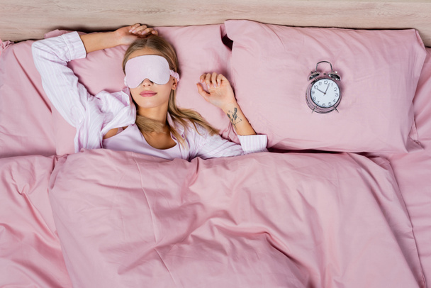 Vista superior de la mujer en la máscara del sueño y pijama acostado cerca del despertador en la cama  - Foto, imagen