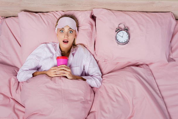 Draufsicht der erstaunten Frau in Schlafmaske mit Pappbecher in der Nähe des Weckers auf dem Bett  - Foto, Bild