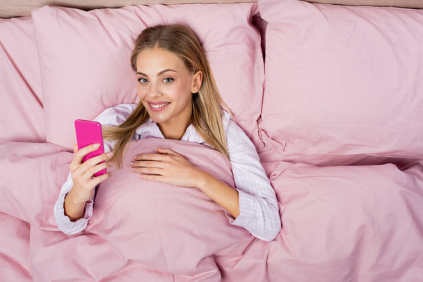 Top view of smiling woman using pink smartphone on bed  - Foto, imagen