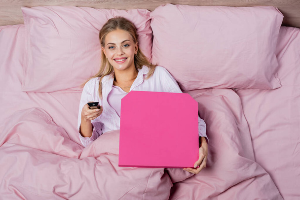 Pohled na usmívající se ženu v pyžamu držící dálkový ovladač a krabici od pizzy na posteli  - Fotografie, Obrázek