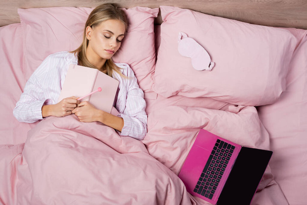 Vista superior de la mujer sosteniendo portátil cerca de la computadora portátil mientras duerme en la cama  - Foto, imagen