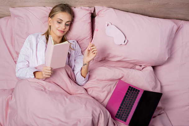 Top view of smiling woman holding notebook and pen near laptop and sleep mask on bed  - Fotoğraf, Görsel