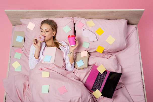 Top view of woman holding coffee to go and donut near books, laptop and sticky notes on bed on pink background  - Photo, Image