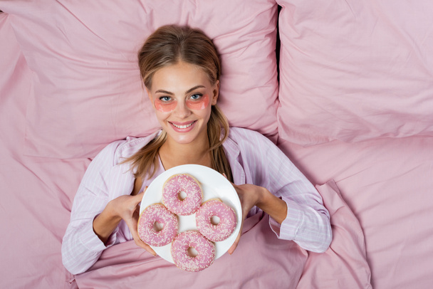 Top view of smiling woman with eye patches holding plate with donuts on bed  - Photo, Image