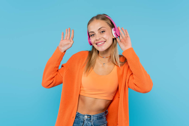 Wesoła blondynka w słuchawkach macha ręką przed kamerą odizolowana na niebiesko  - Zdjęcie, obraz