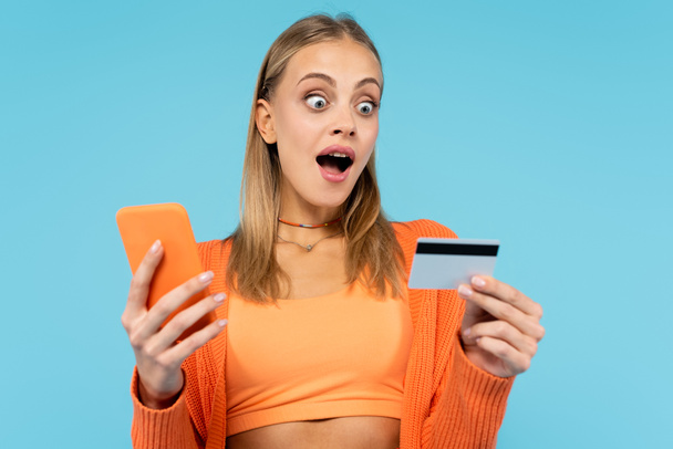 Ενθουσιασμένη ξανθιά γυναίκα που κατέχει πιστωτική κάρτα και smartphone απομονώνονται σε μπλε  - Φωτογραφία, εικόνα