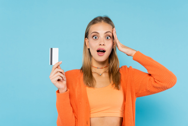 Shocked blonde woman holding credit card isolated on blue  - Фото, зображення