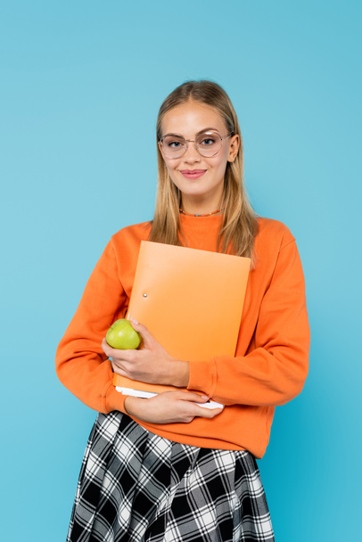Студент блондинки в окулярах тримає паперову теку і яблуко ізольовано на синьому
  - Фото, зображення